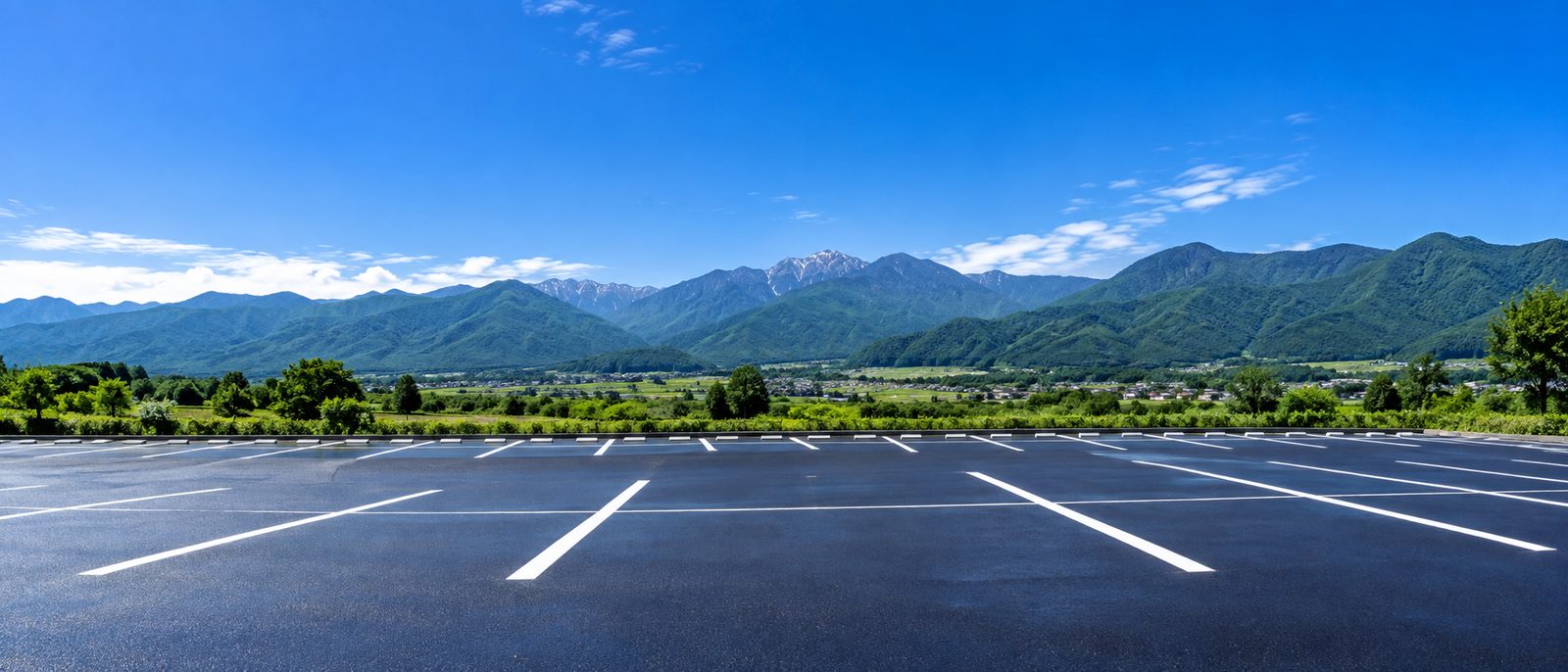 舗装を終えたばかりの黒く艶のあるアスファルトの駐車場と白線、奥に信州の山並みと青空のイメージ